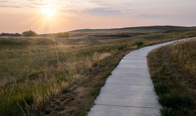 Concrete trail curves gradually to right. Orange and pink sunrise and rolling hills to left.