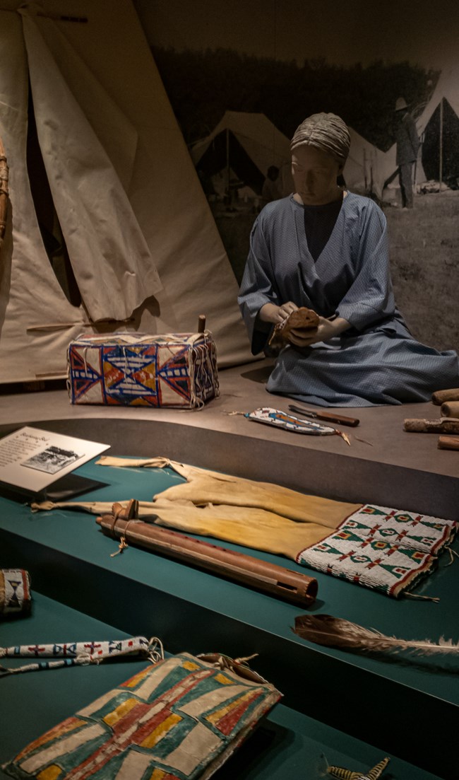 A mannequin of woman in a blue dress works next to a tipi. Native American artifacts made of leather, wood, and colorful beads surround her in the foreground.