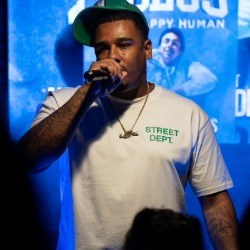 Black man in white t-shirt and green baseball cap speaks into a microphone in front of a crowd.