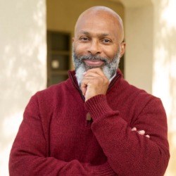 Smiling Black man with salt and pepper beard, bald head, and red sweater looks directly at camera.