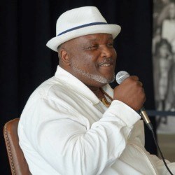 Black man in white fedora and button-down holds microphone and smiles at the camera.
