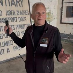 White man in black vest and turtleneck gestures with one hand and talks to group off-camera.