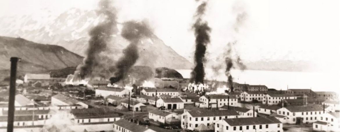 a seaside town with numerous buildings on fire and throwing smoke up into the air.