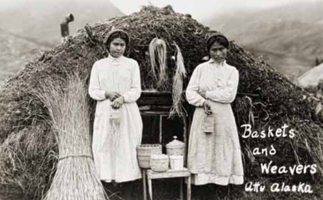 two young women stand in front of a sod house holding grass baskets.