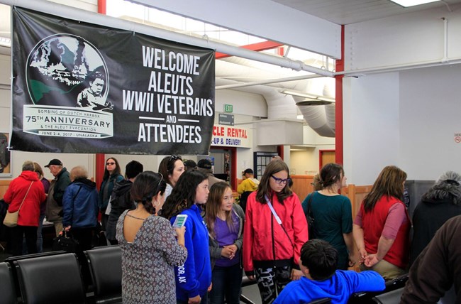 People gather under a banner reading "Welcome Aleuts WWII Veterans and Attendees"