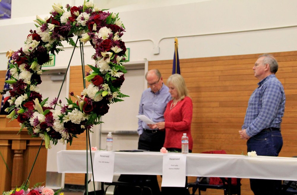 A floral wreath on a stand with three people in the background.