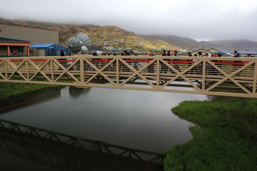 A group of people cross a bridge over calm water with hills in the background