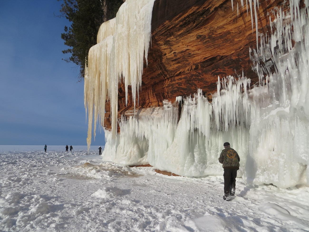 Sunny day with a person walking in front of icy cliffs with more people in the distance on the ice.