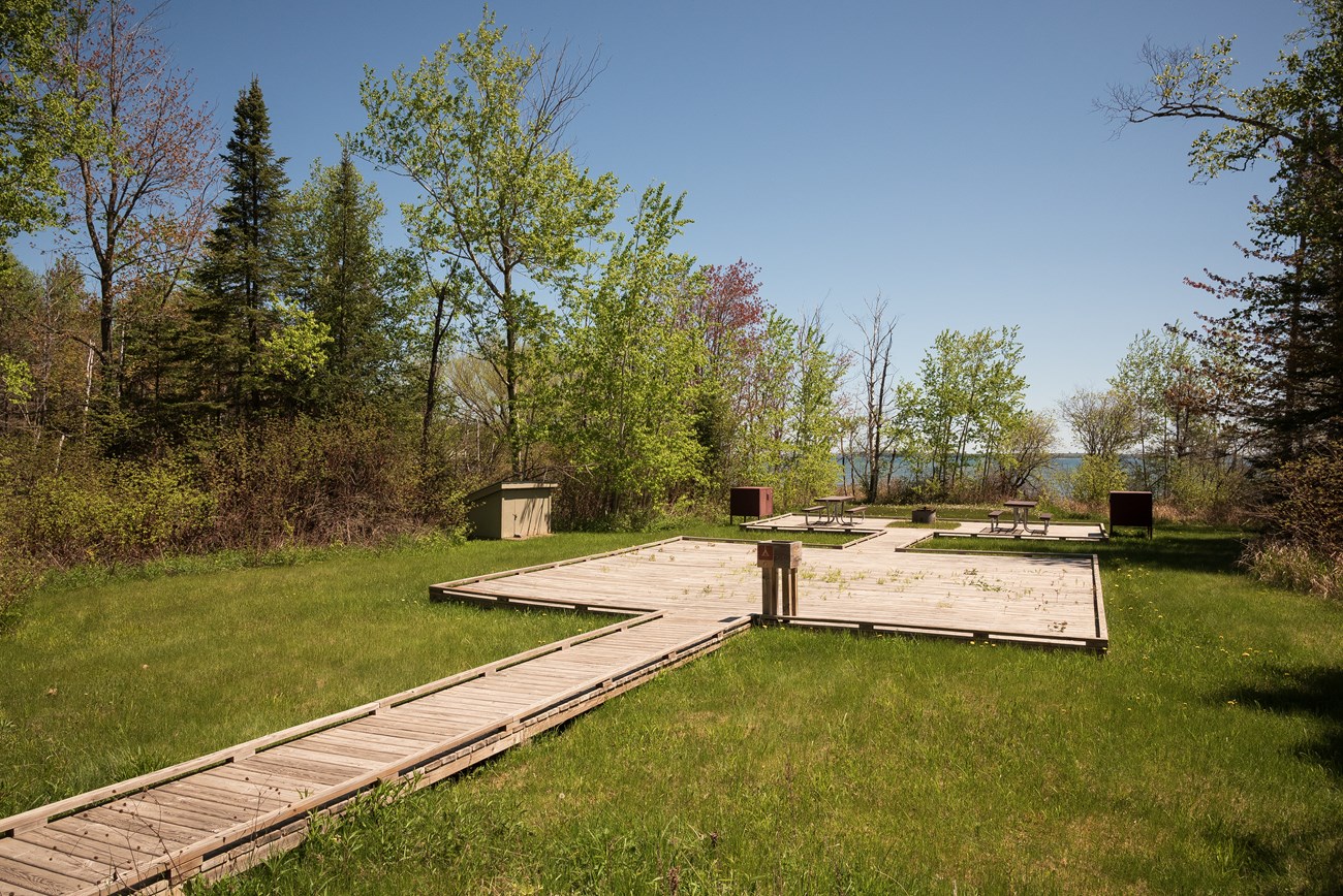 Photograph of a grassy area with wooden walkway leading to a picnic tables on wooden platforms. Trees and a lake are visible in the background on a clear, sunny day.