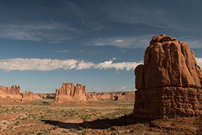 Expanse view of a landscape of red sand and soil, dotted with green vegetation. In the foreground a massive red rock protrudes from the ground. In the distance are more large red rocks with a blue sky and a thin line of white clouds.