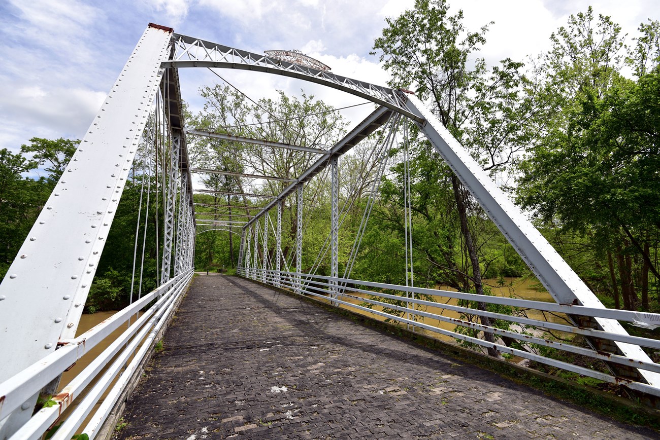 View down a rusting white metal truss bridge with a wooden floor spanning a muddy river.