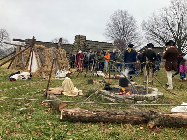 Living historians talk to visitors in front of a campfire near log huts
