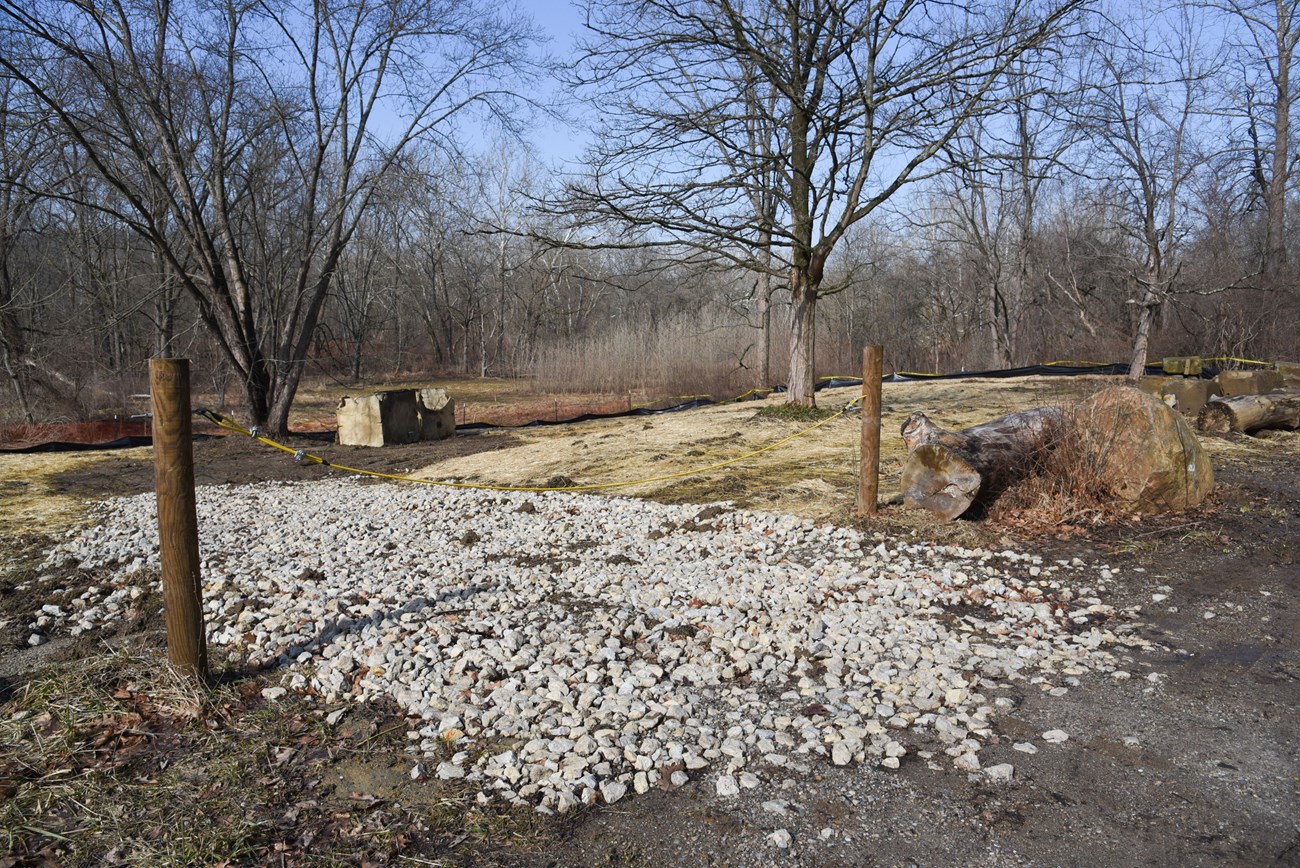 A cable gate blocks access across a white gravel driveway; the ground beyond the gate is covered in straw with trees in the distance.