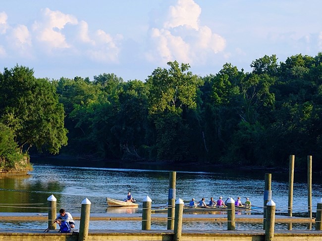 A man fishes off a dock along a river. Several people sit in a large kayak. Behind them a densely forested shoreline is silhouetted by a blue sky and large, white clouds.