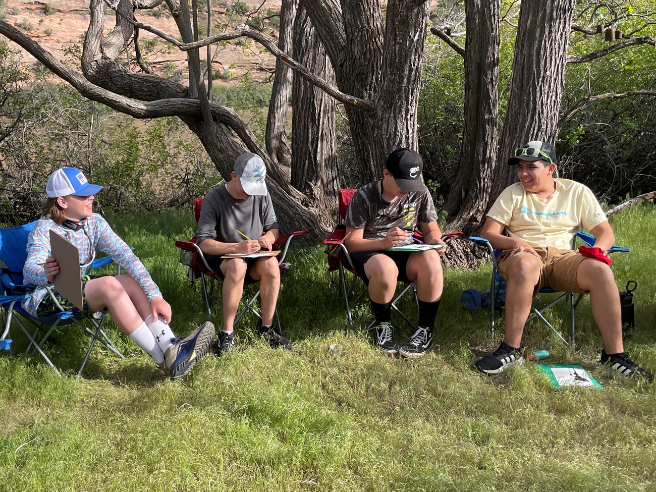 Four students sitting in camp chairs with clipboards and pencils. The two on either end share smiles over their work, as the two in the middle are focused on their writing or drawing.