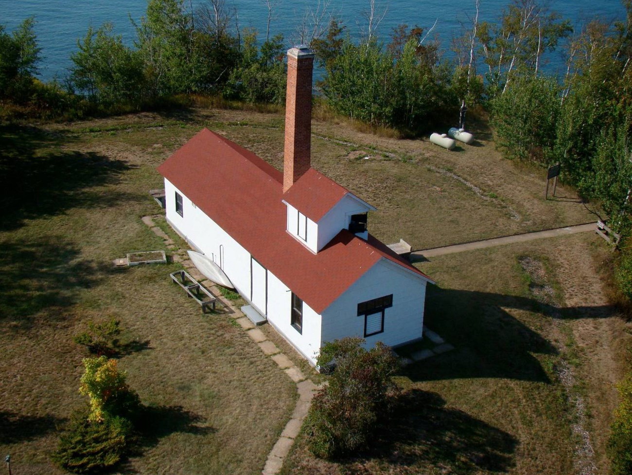 A long, one story building with a brick chimney is surrounded by a landscape of concrete pathways, turf, tree-covered embankment, and water beyond.