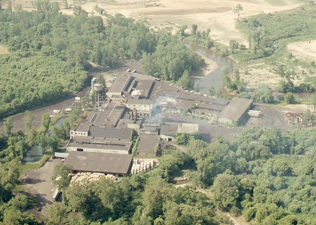 A water tower overlooks a tight cluster of factory buildings beside a river bend and forest.