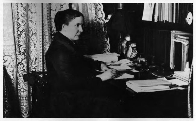 A woman sits at a desk strewn with papers. LOC