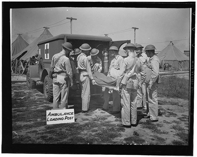 A group of uniformed soldiers practice loading an injured person on a stretcher into the back of an ambulance. A military camp is visible in the background.