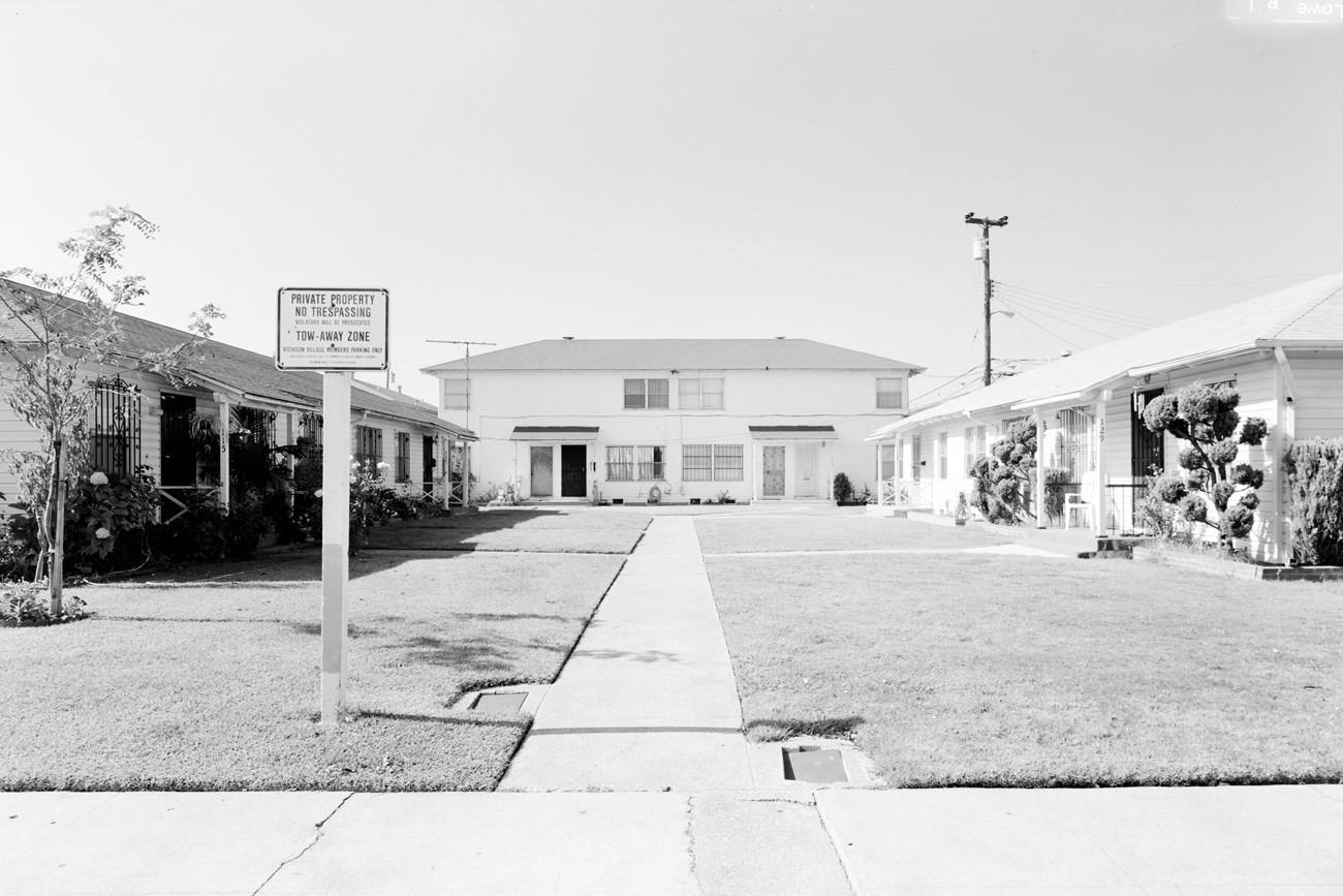 Modest, white clapboard single story homes faced central grassy courtyard in planned neighborhood of government housing. Sing reads “private property no trespassing.”