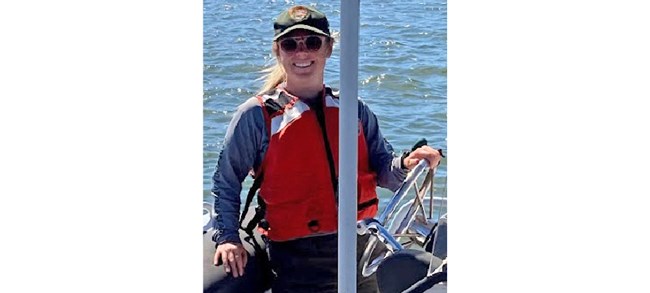 Button in a NPS ball cap, orange life vest, and sunglasses, smiling at the helm of a boat.