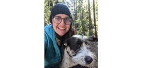 Woman in blue jacket with blue knit hat holds a black and wither terrier in front of a stand of evergreen trees