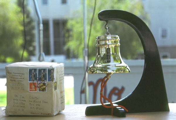 Image of small bell hanging from curved black stand, a small package is at left of the bell.