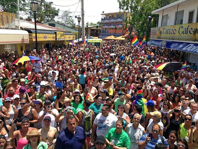 City street lined with businesses and densely packed with crowds of people. Gay pride flags and other rainbow items protrude from the crowd.