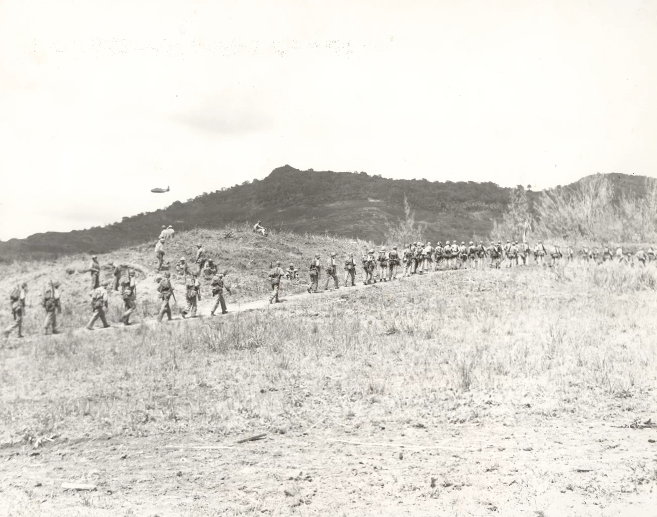 Members of the 2nd Battalion, 4th Marines move into position on the front lines of the foothills of Mount Alifan, home of Colonel Tsunetaro Suenaga's command post. Soldiers march in a single file across a barren landscape, with rugged hills in the background. A plane flies low in the background.