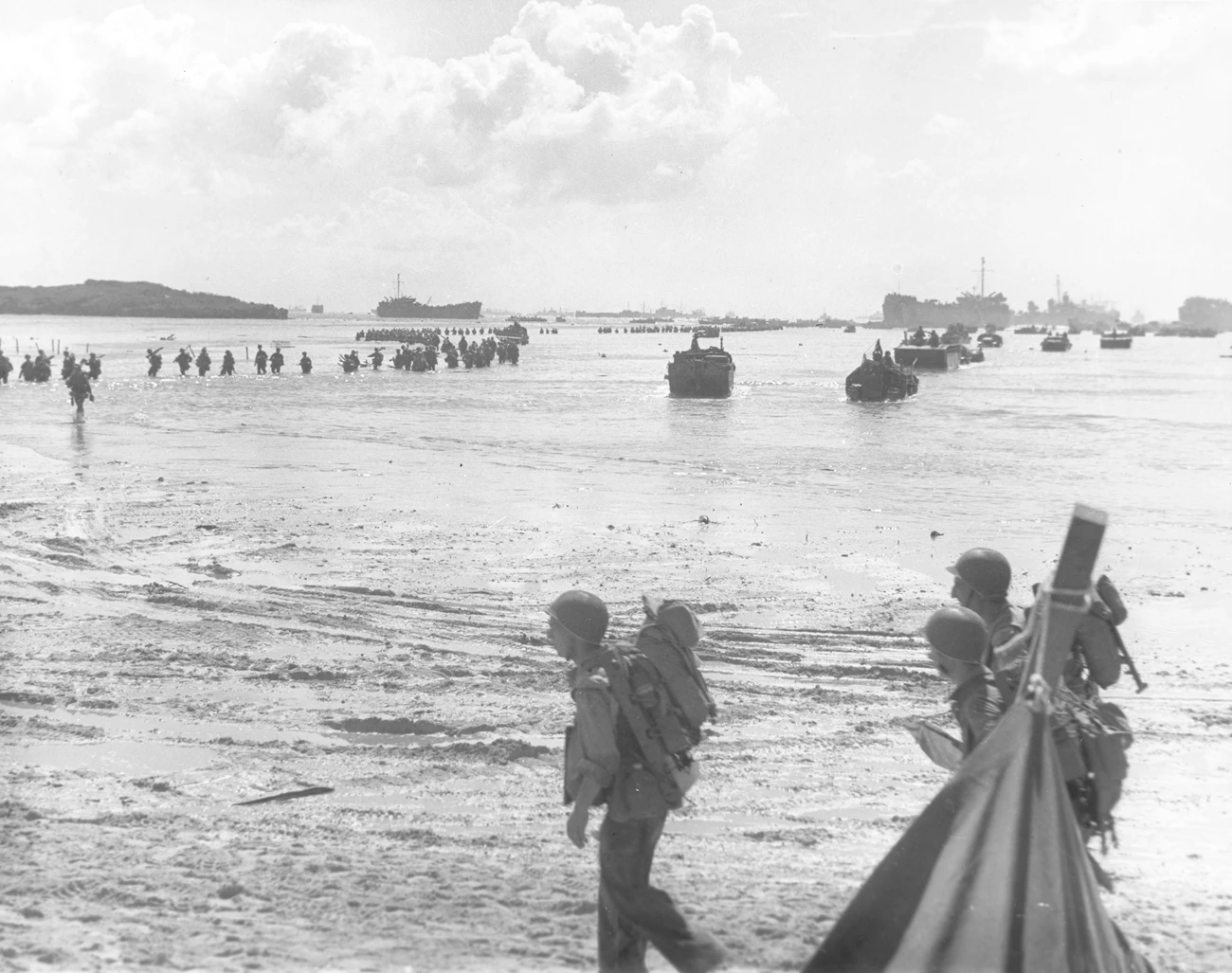 Members of the 306th Regiment, 77th Infantry Division wade ashore on Hågat Beach during the Battle of Guam. There were no Amtracs to transport the soldiers to shore, so they waded the last 400 yards over the reef. Soldiers wade through shallow waters toward the beach, with landing boats and ships in the background under a cloudy sky.