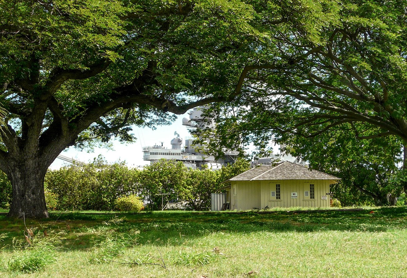 Bungalow 90 with USS Missouri in the background. Small yellow building in grassy field dwarfed by WWII battleship in background.