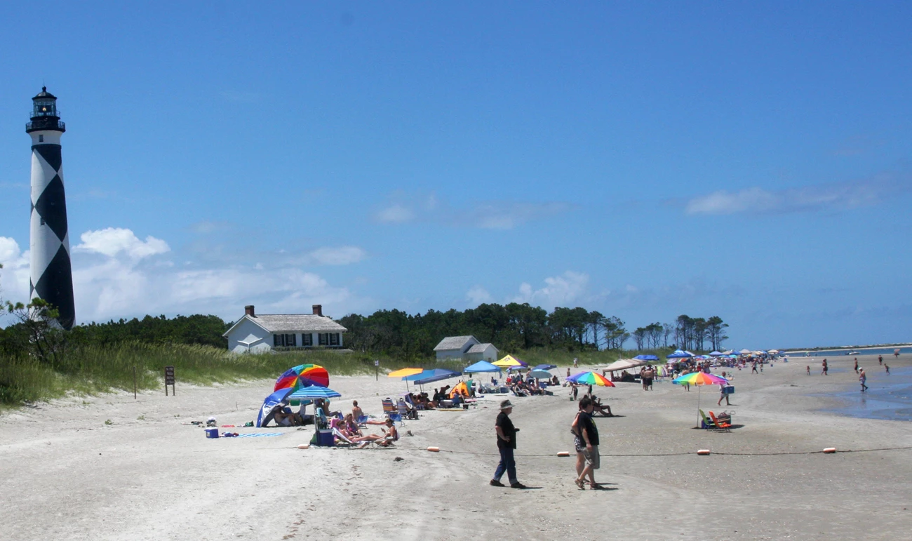 Cape Lookout lighthouse beach A sunny beach with a black and white diamond-patterned lighthouse on the left and many people under colorful umbrellas.