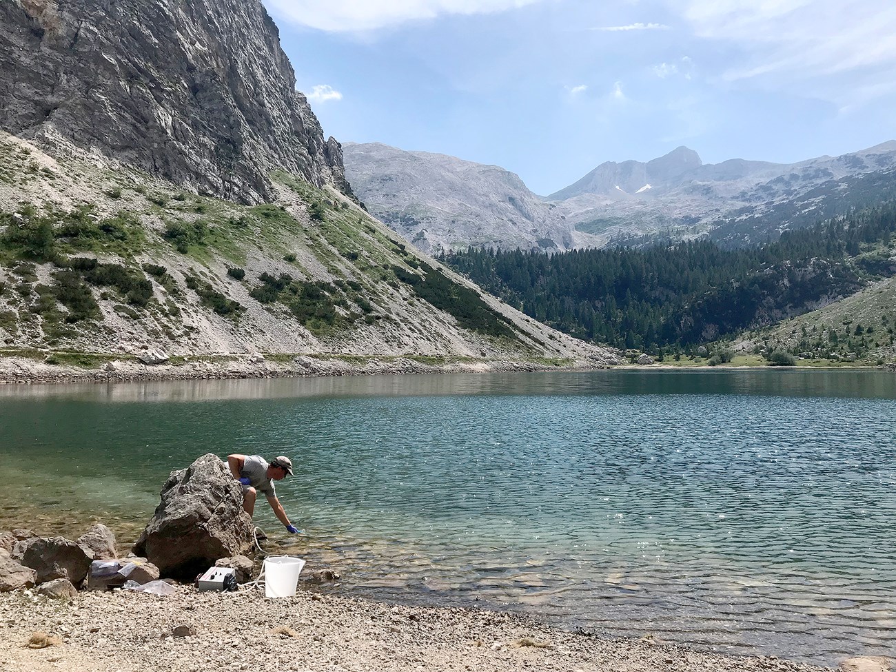 A man in a white shirt and baseball cap dips a sampling cup into a blue lake. he is surrounded by mountains, trees, and blue sky.