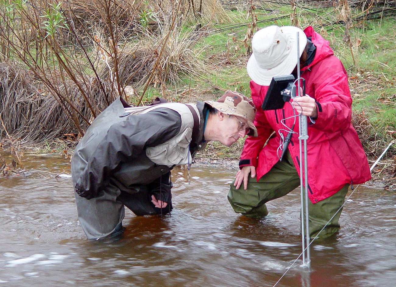 Collecting stream gauge data A man bending over to examine an instrument held by a woman. Both are standing in a stream.