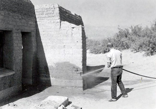 Man uses hose and blasts wall of adobe building with water.