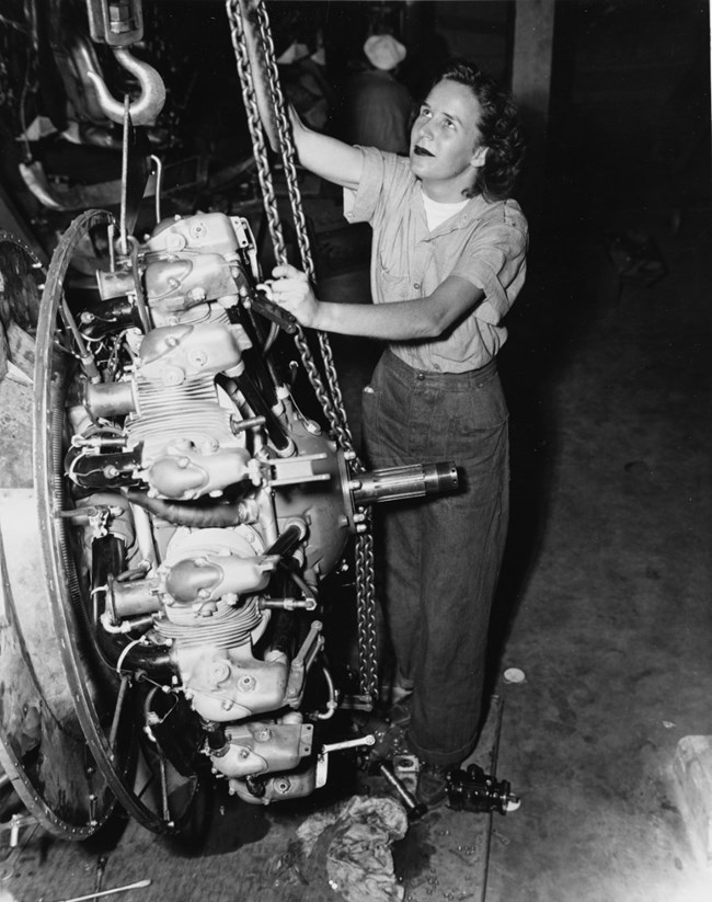 Black and white photo of a woman in button down and pants pulling a chain attached to an engine