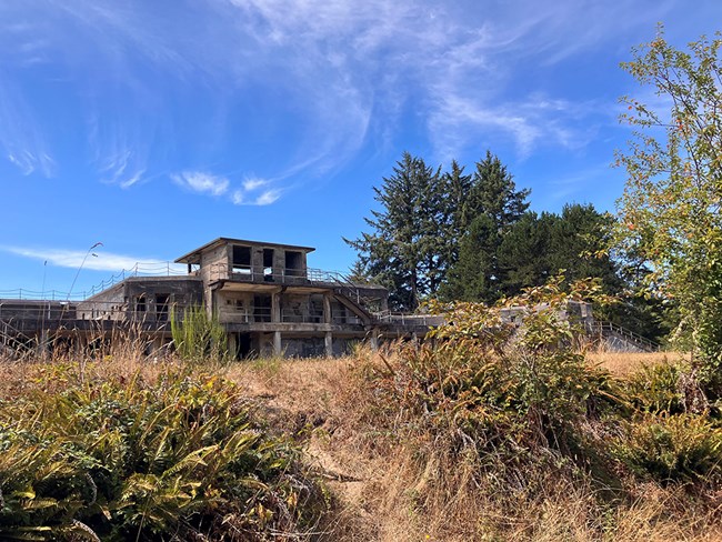 A color photograph looking up a small rise of grasses at the façade of Battery Russel. It is a grey, boxy structure built of concrete. No windows or doors exist anymore. The sky above is bright blue with wisps of cloud.