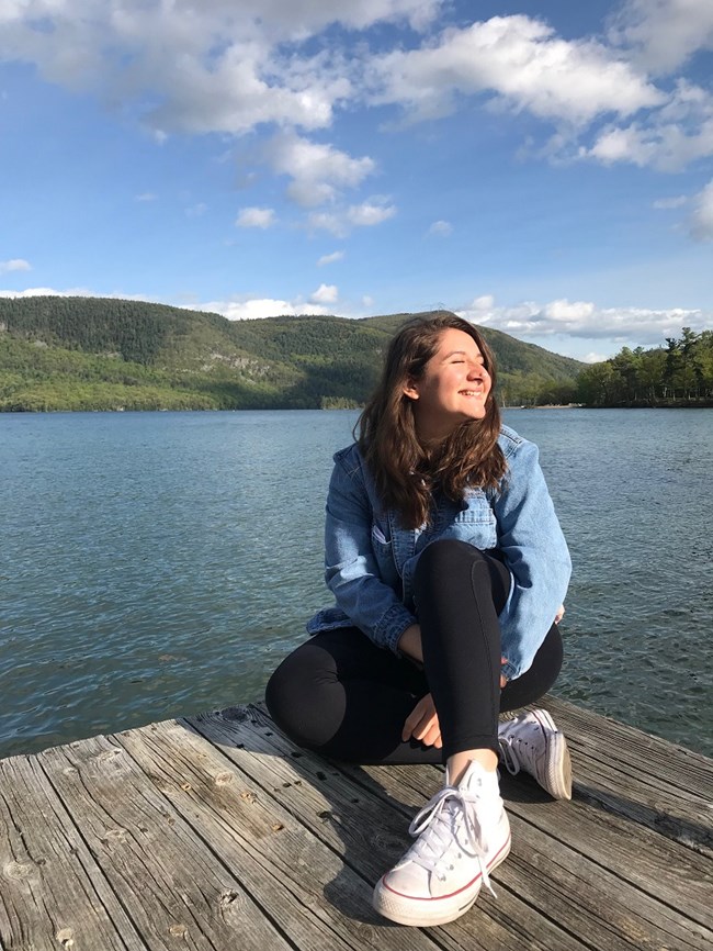 evelyn sitting on a wooden structure with a lake behind her, smiling while catching some sunrays