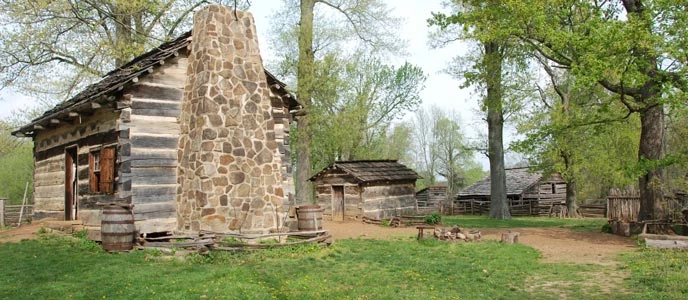 Log cabin with stone chimney with log smokehouse and log barn in the background. Farm yard and cabin