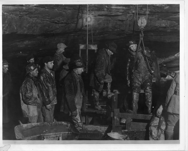 A black and white photo of a group pf men looking on as another man is lowered down on a rope.