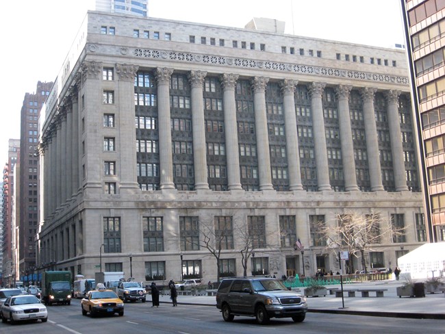 Eleven-story light-colored building split into three sections. The bottom floors feature smooth exterior walls and thirteen-bays. Middle section features twelve Corinthian columns. Top section consists of the cornice and roofline.