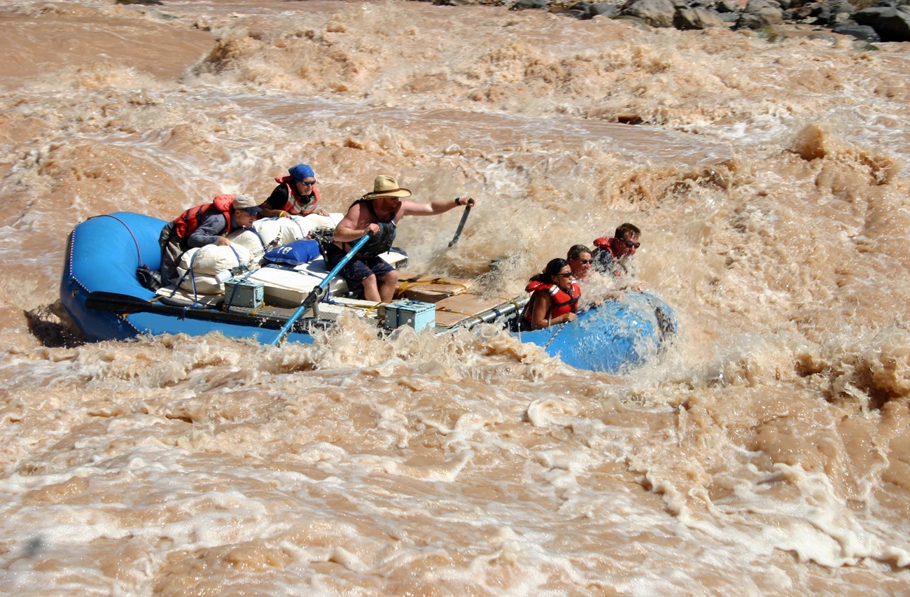 Whitewater rafting Six people and a lot of gear in a big, blue raft in the midst of roiling river rapids. One is paddling as the others hold on to ropes in the front or back of the raft. The fast-moving water is the color of coffee with cream.