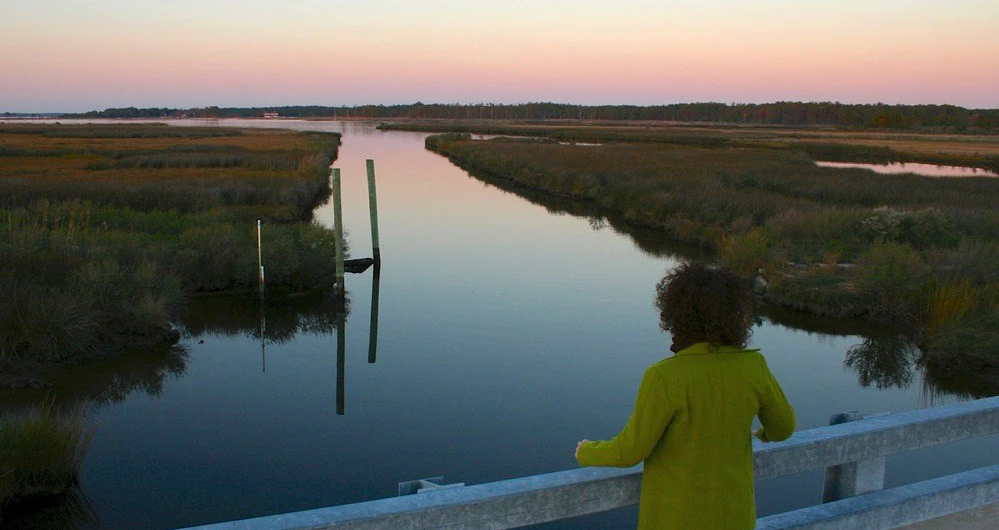 Stewart's Canal in Eastern Maryland a woman along a low bridge gazes out at a grassy wetland with canals as the sun begins to set