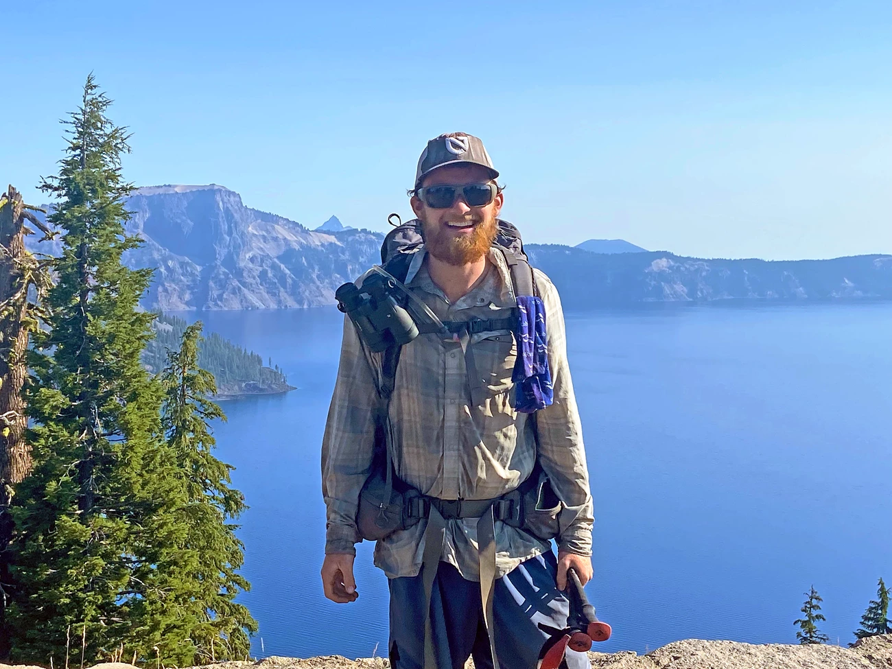 Will Babb at Crater Lake The author, with sunglasses and a beard, smiles for a photo in front of Crater Lake, which appears faded in haze from wildfire smoke. He carries a large backpacking pack, binoculars, and a pair of hicking poles.