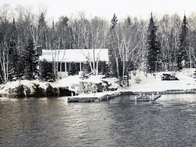 cabin with a snow covered roof sits on the snowy banks of Rock Harbor