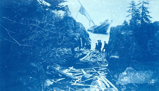 SS ALGOMA barrels and boards from SS Algoma are strewn between a gap in the outer rocks of Mott Island, ship sinking in the background, people looking on