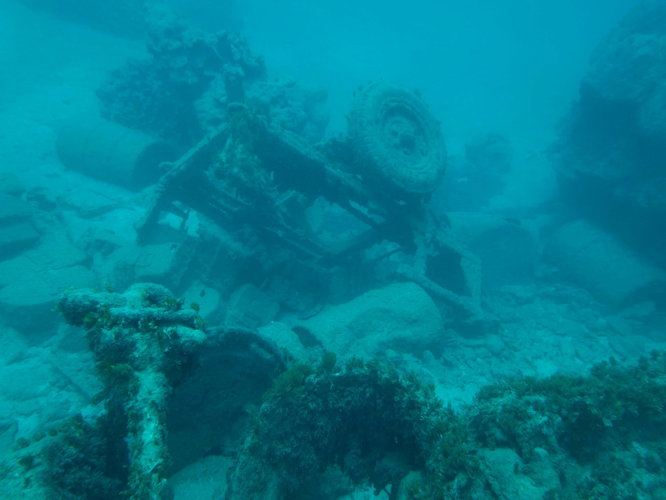 A World War II-era jeep trailer loaded with vehicle parts and equipment offshore at Hågat Beach. It was most likely a casualty of the 77th Division's disorganized landing. Underwater image of a rusty, algae-covered vehicle wreck, surrounded by scattered debris and rocks.