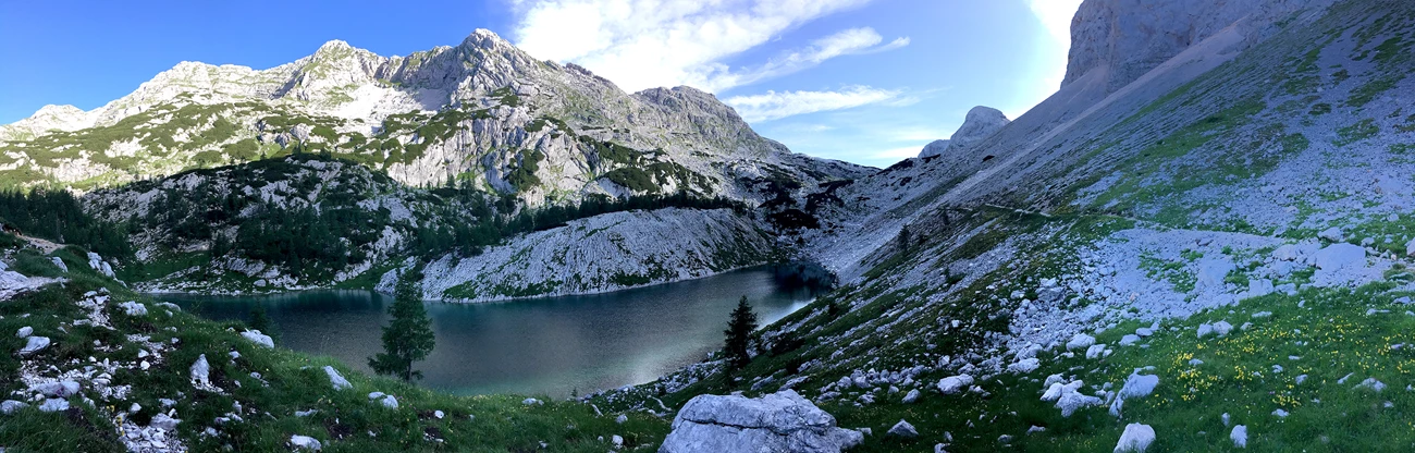 Jezero v Ledvicah, or "Lake at Ledvicah" A panoramic image of a blue-green lake surrounded by rocky gray mountains, green meadows, green trees, and blue skies.