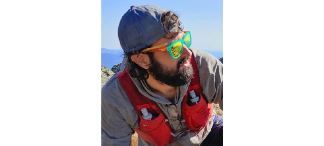Tourville outside in the mountains with a backwards ball cap, colorful sunglasses, and a bright red vest with waterbottles in the front pockets.