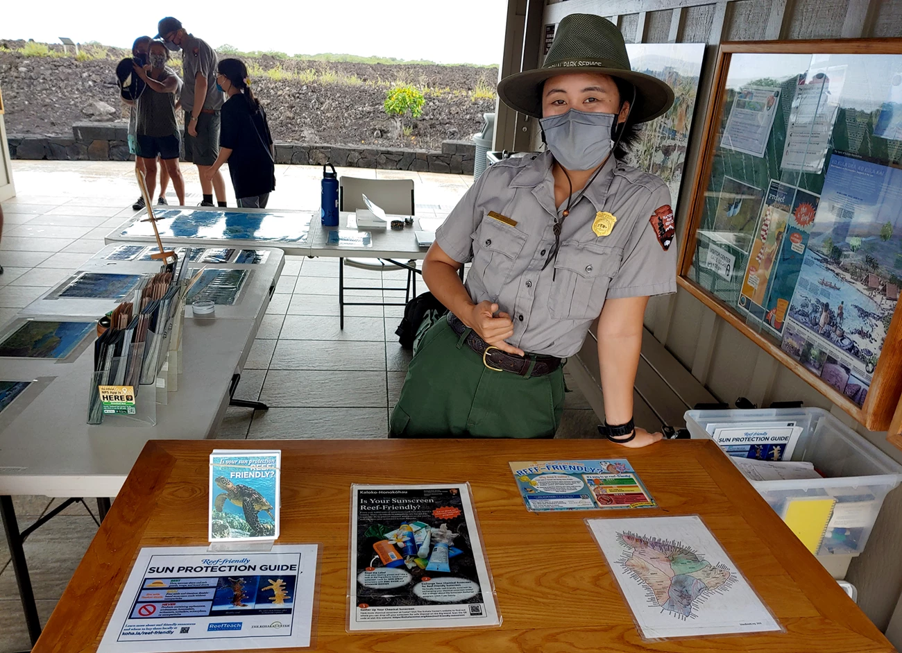 Sunscreen information booth A woman in an NPS uniform standing behind a table with sunscreen information on it. In the background is another ranger and three visitors.